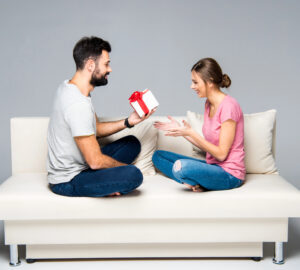 A man exchanging a gift with a woman seated on a white couch.