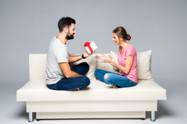 A man exchanging a gift with a woman seated on a white couch.